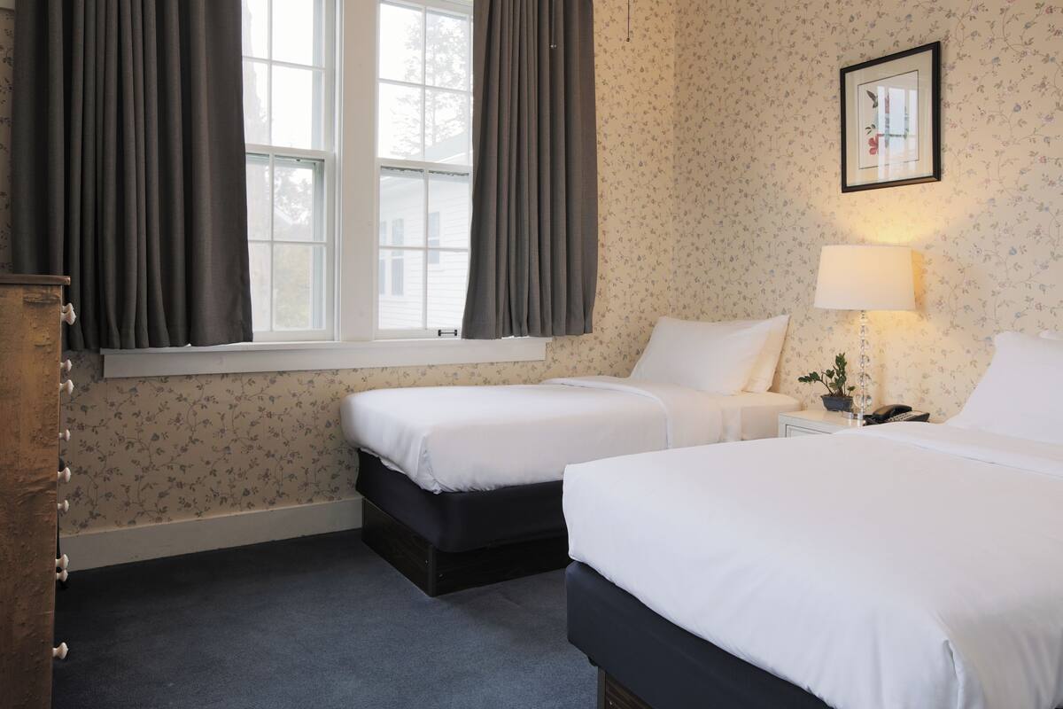 A hotel room with two beds, a window with curtains, a bedside lamp, and a floral wallpaper.