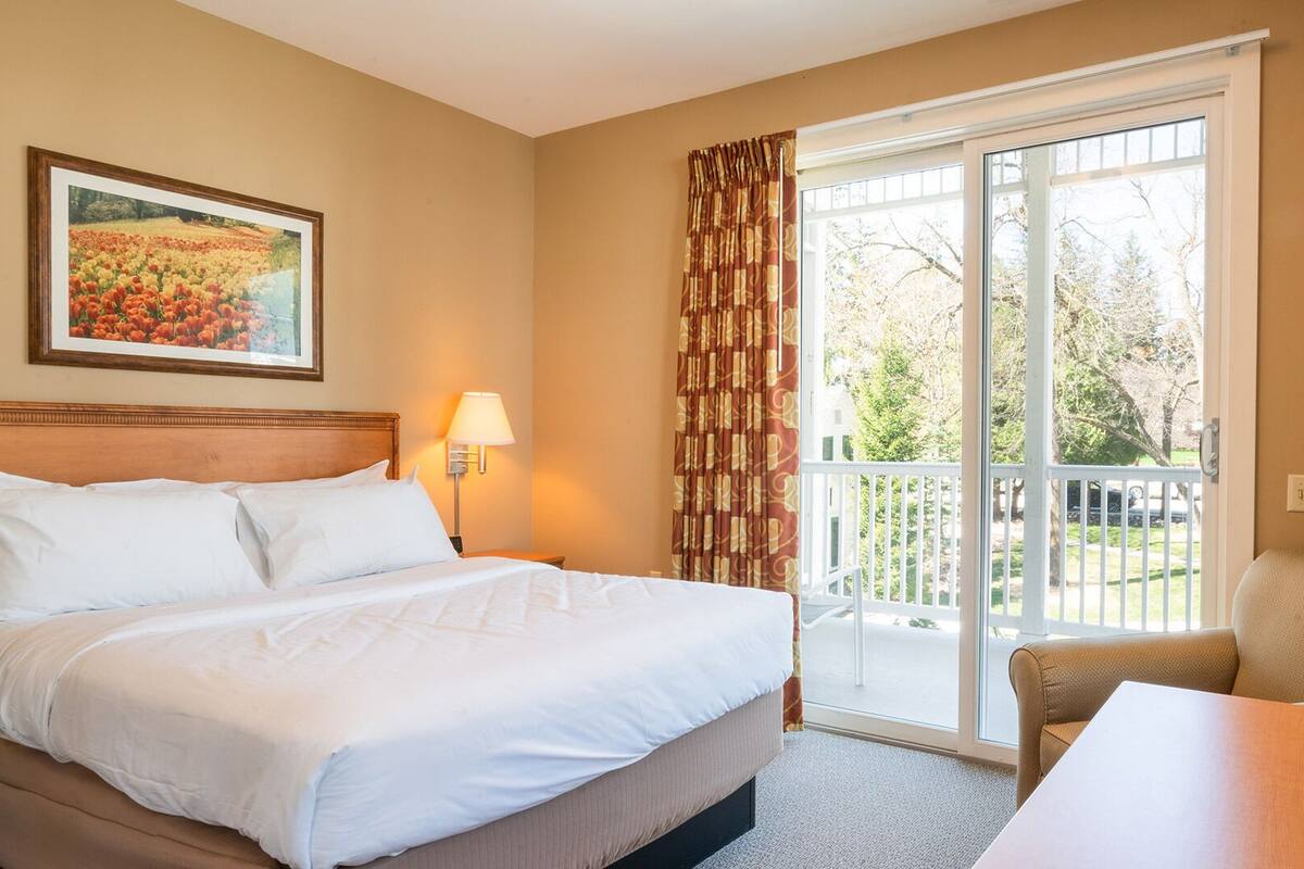A hotel room with a bed, a chair, a table, a lamp, a painting, and a balcony with curtains.
