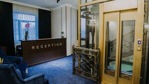 Ein Empfangsbereich mit einer Rezeption, auf der „RECEPTION“ steht, einem blauen Teppich, einem blauen Stuhl und einem gläsernen Aufzug mit goldener Umrandung.