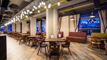 A spacious restaurant with wooden tables and chairs, hanging light fixtures, and large windows with curtains.