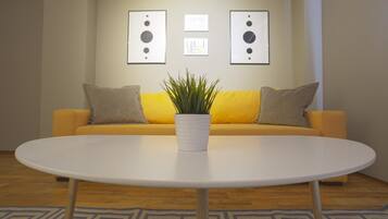 A room with a yellow sofa, a white table, and a potted plant. There are framed pictures on the wall.