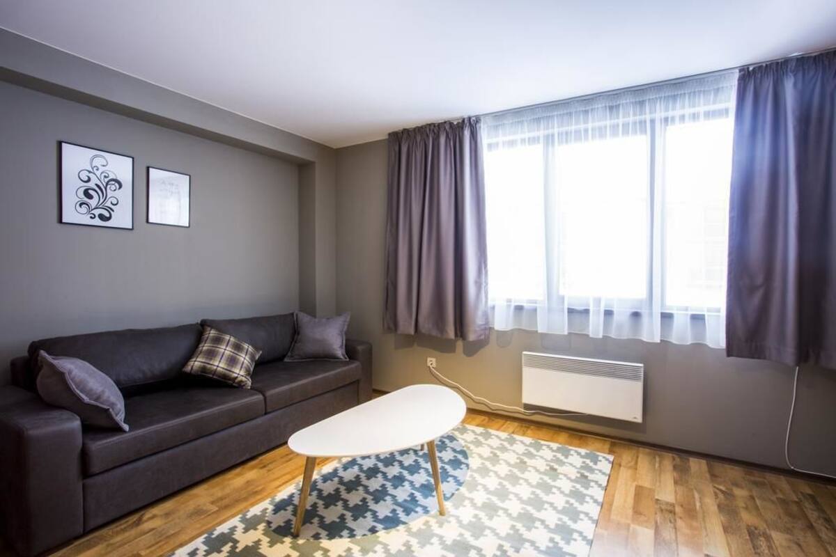 Ein modernes Wohnzimmer mit einem dunkelbraunen Sofa, einem weißen Couchtisch und einem gemusterten Teppich. An der Wand hängen zwei gerahmte Bilder, ein Fenster mit Vorhängen und ein Heizkörper.