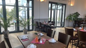 Een eetgedeelte met tafels voor vier personen, potplanten en grote ramen met uitzicht op de straat.