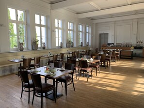 A spacious dining area with multiple tables and chairs, large windows, and a kitchen in the background. - Parkhotel Putbus(Adults Only) (Putbus)