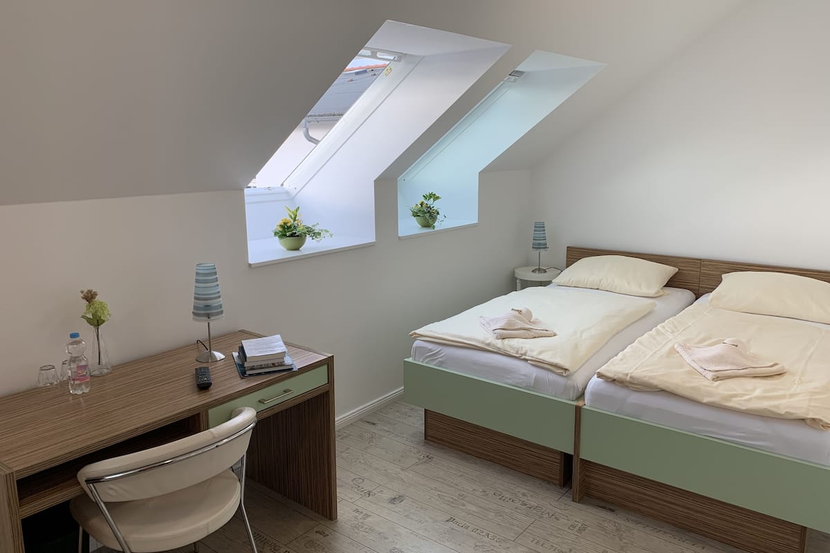 Un dormitorio con techo inclinado, un escritorio, una silla, dos camas y una ventana pequeña con una planta.