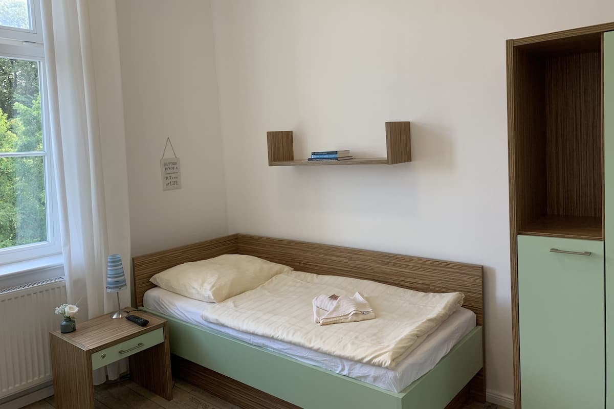 Una cama bien hecha con un cojín y una manta doblada, una mesita de noche con una lámpara y un control remoto, un pequeño estante con libros y un armario.