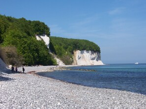Een kiezelstrand met een witte klif en begroeiing, een boot in het water, en mensen die langs de kust wandelen.