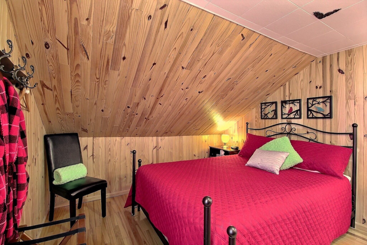 Un dormitorio con techo inclinado, paredes de madera, cama con ropa de cama roja, una silla con cojín verde y obras de arte en la pared.