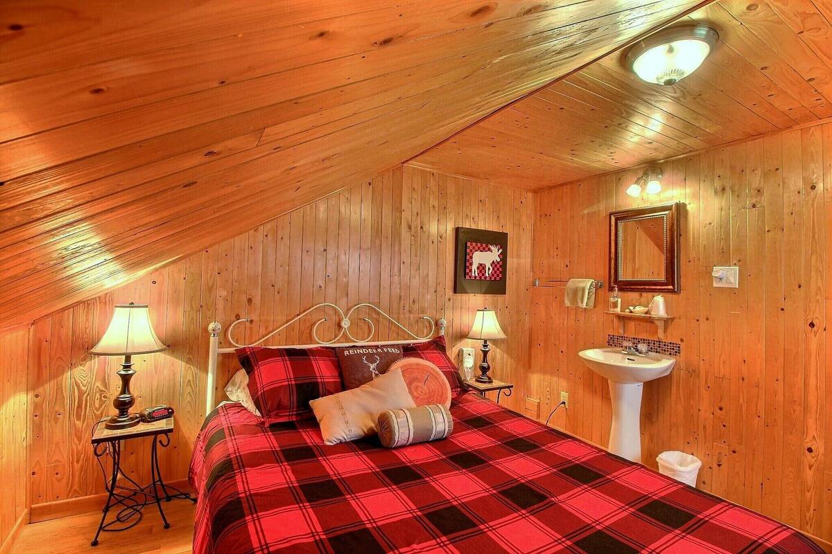 Un dormitorio acogedor con techo de madera, una cama con colcha a cuadros rojos, una mesita de noche con lámpara, un espejo y un lavamanos.
