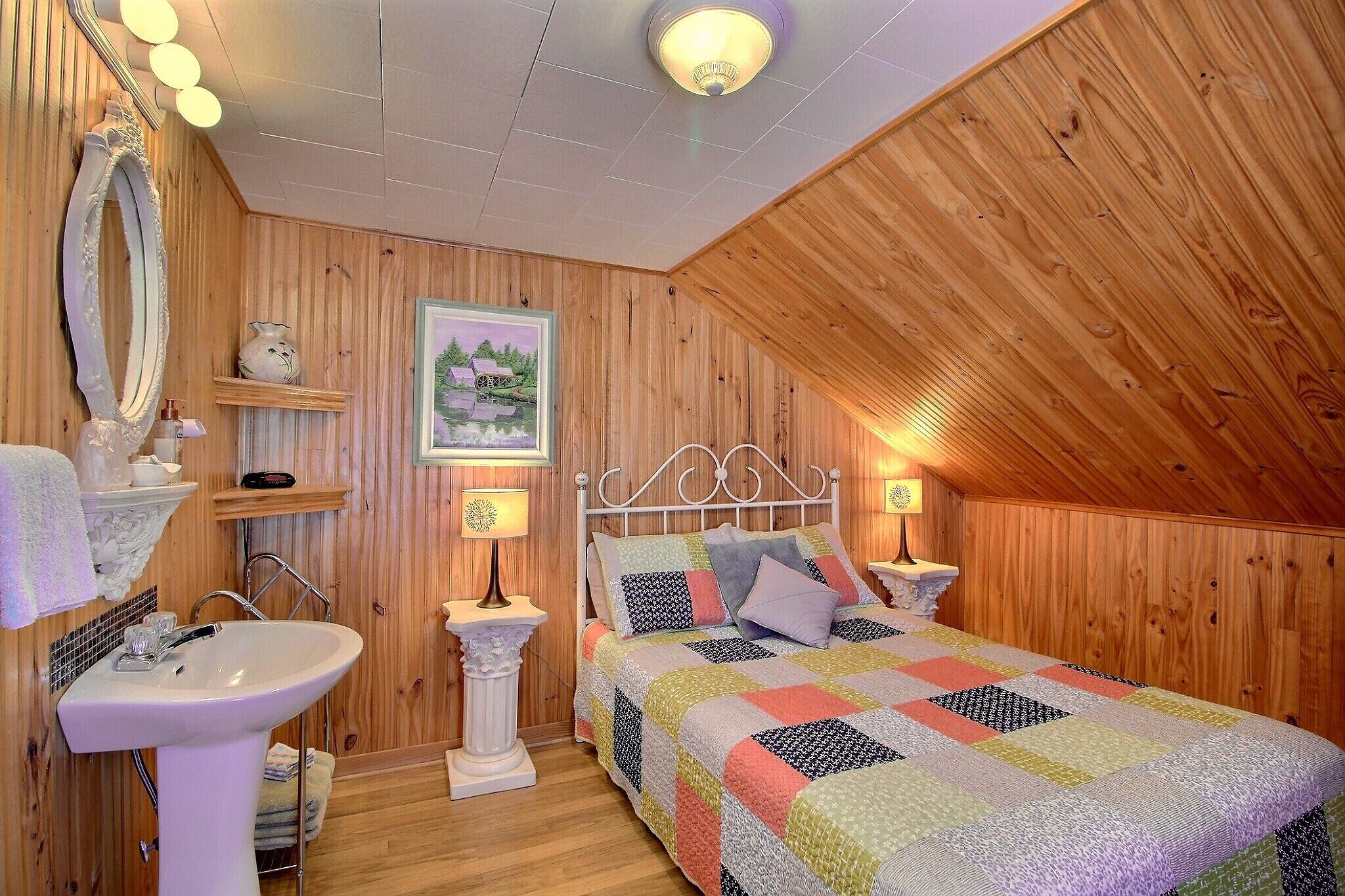 Une chambre à coucher chaleureuse avec des murs en bois, un lit recouvert d’une courtepointe colorée, une table de chevet blanche, une lampe blanche et un miroir.