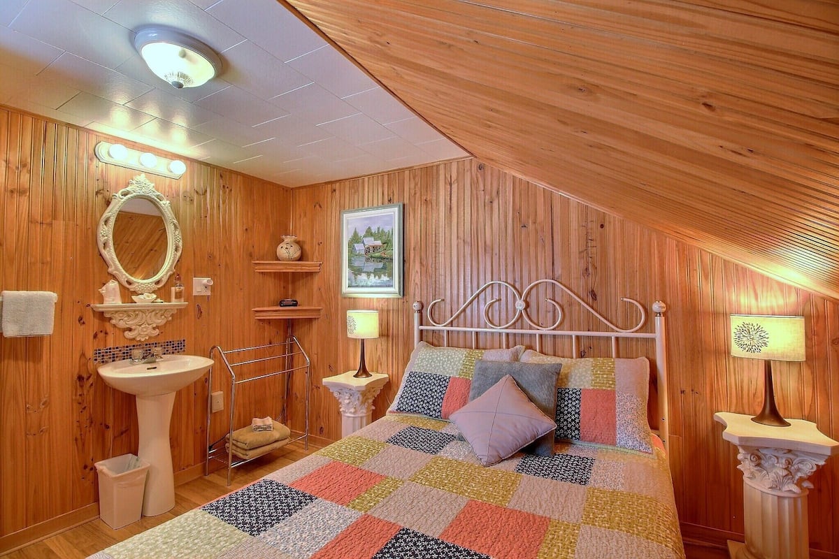 Un dormitorio acogedor con techo de madera, una cama con colcha colorida, cabecera de metal blanco, lámpara de noche, un espejo y un pequeño estante con objetos decorativos.