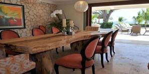 A dining area with a large wooden table, red upholstered chairs, a stone wall, and a view of the outdoors.