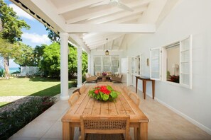 A covered patio with a wooden table and chairs, surrounded by white walls and a view of the garden. - Wag Water Villa, Ocho Rios 3BR (Ocho Rios)