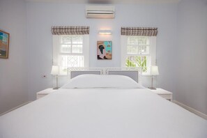 A bedroom with a large bed, two bedside tables with lamps, a wall-mounted air conditioner, and two windows with curtains.