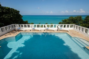 A pool with a view of the ocean, surrounded by white railings and lounge chairs.