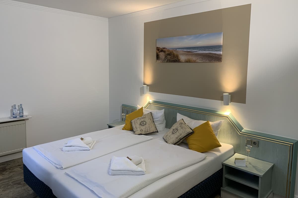 A hotel room with a bed, bedside tables, a nightstand, a wall-mounted painting, and a small shelf with a glass.