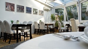 A dining area with a table set for four, surrounded by white chairs, a large window with a view of greenery, and artwork on the walls. - Hotel Residenz Lauterbach  (Putbus)