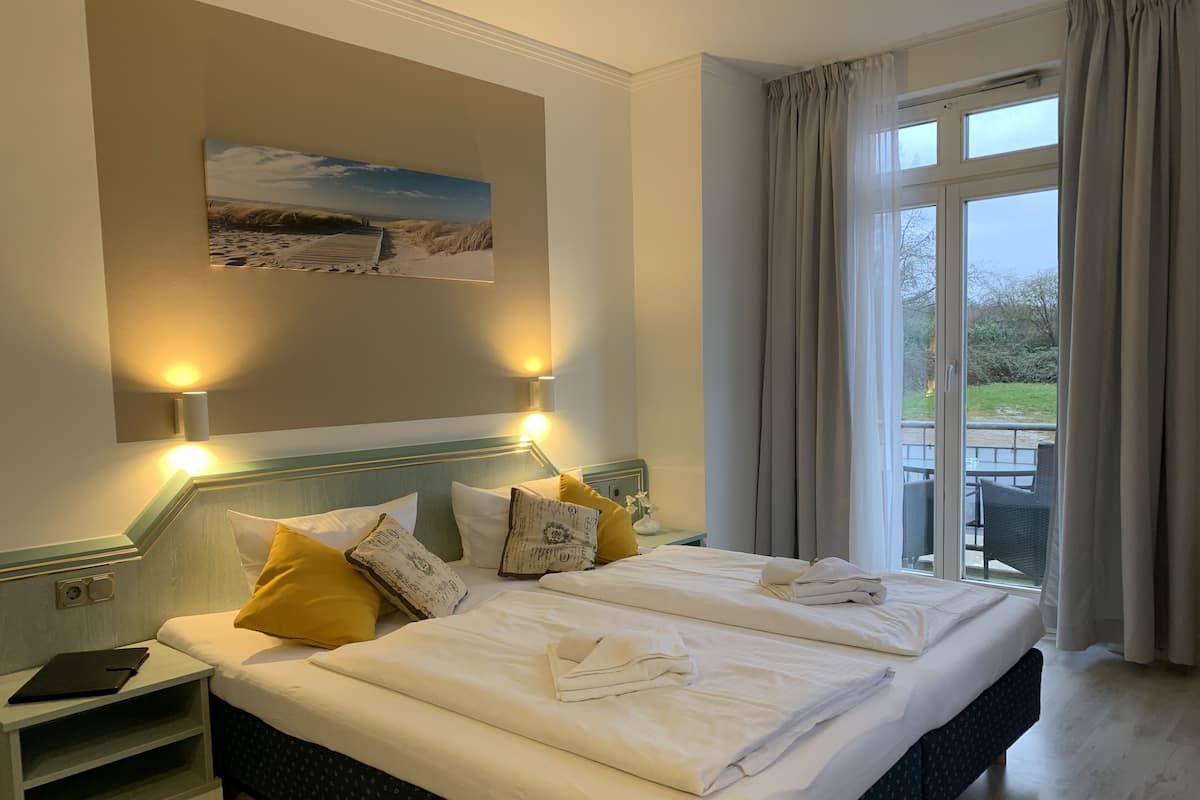 A neatly made double bed with white linens, a headboard with wall-mounted lights, a bedside table with a book, and a view of the outdoors through a large window with curtains.