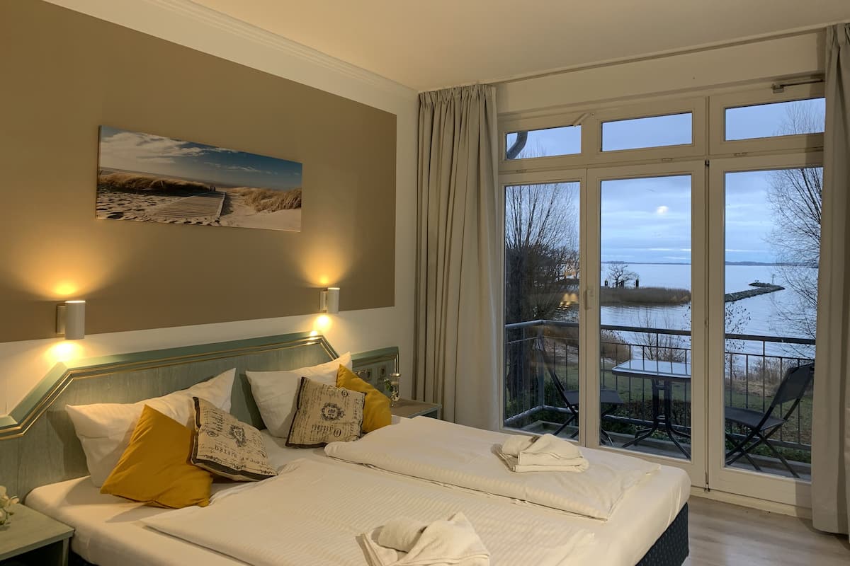 A hotel room with a large bed, a painting on the wall, and a view of a body of water through the windows.