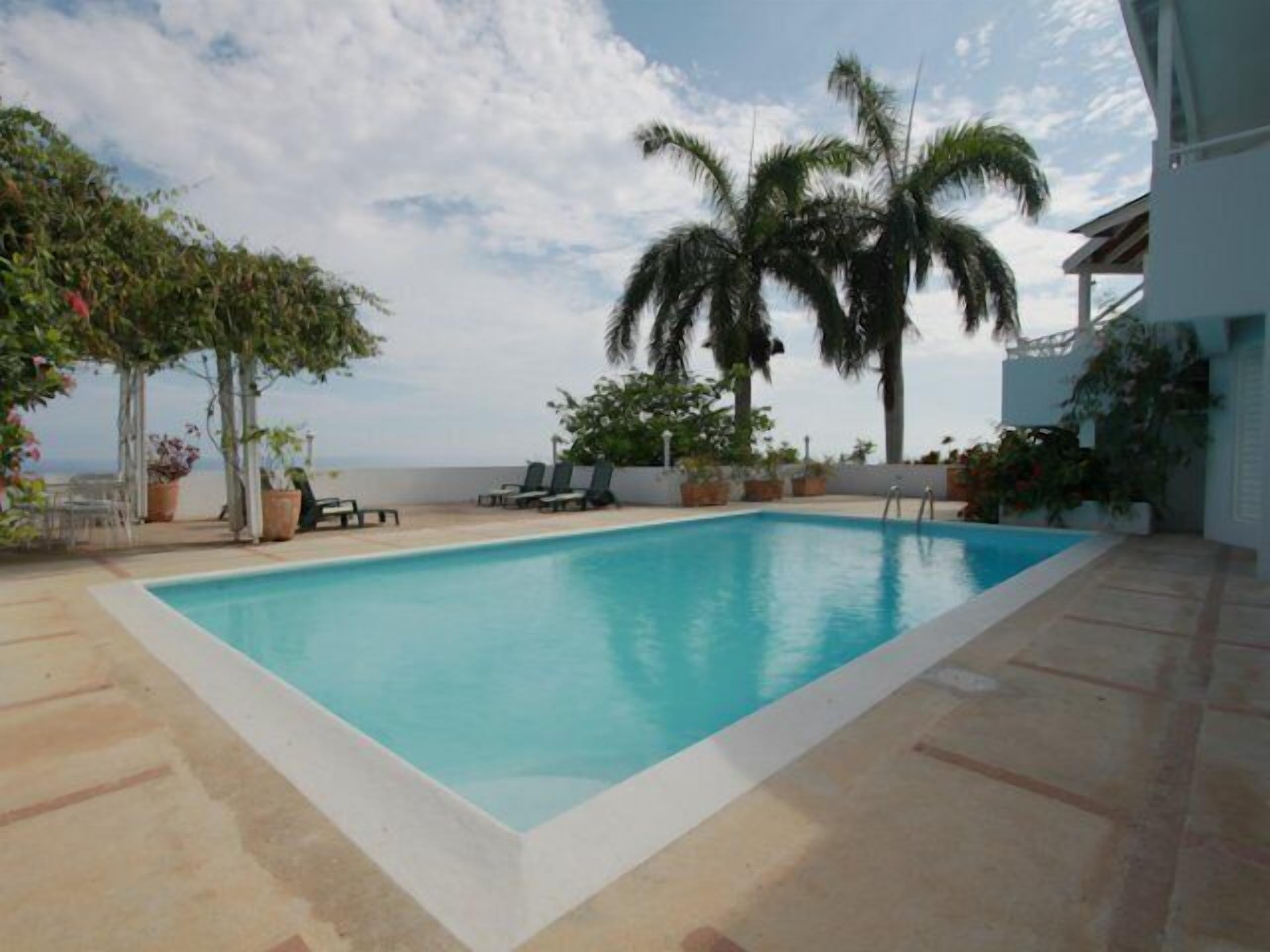 A rectangular swimming pool with a white edge, surrounded by a tiled deck. There are lounge chairs and potted plants around the pool.