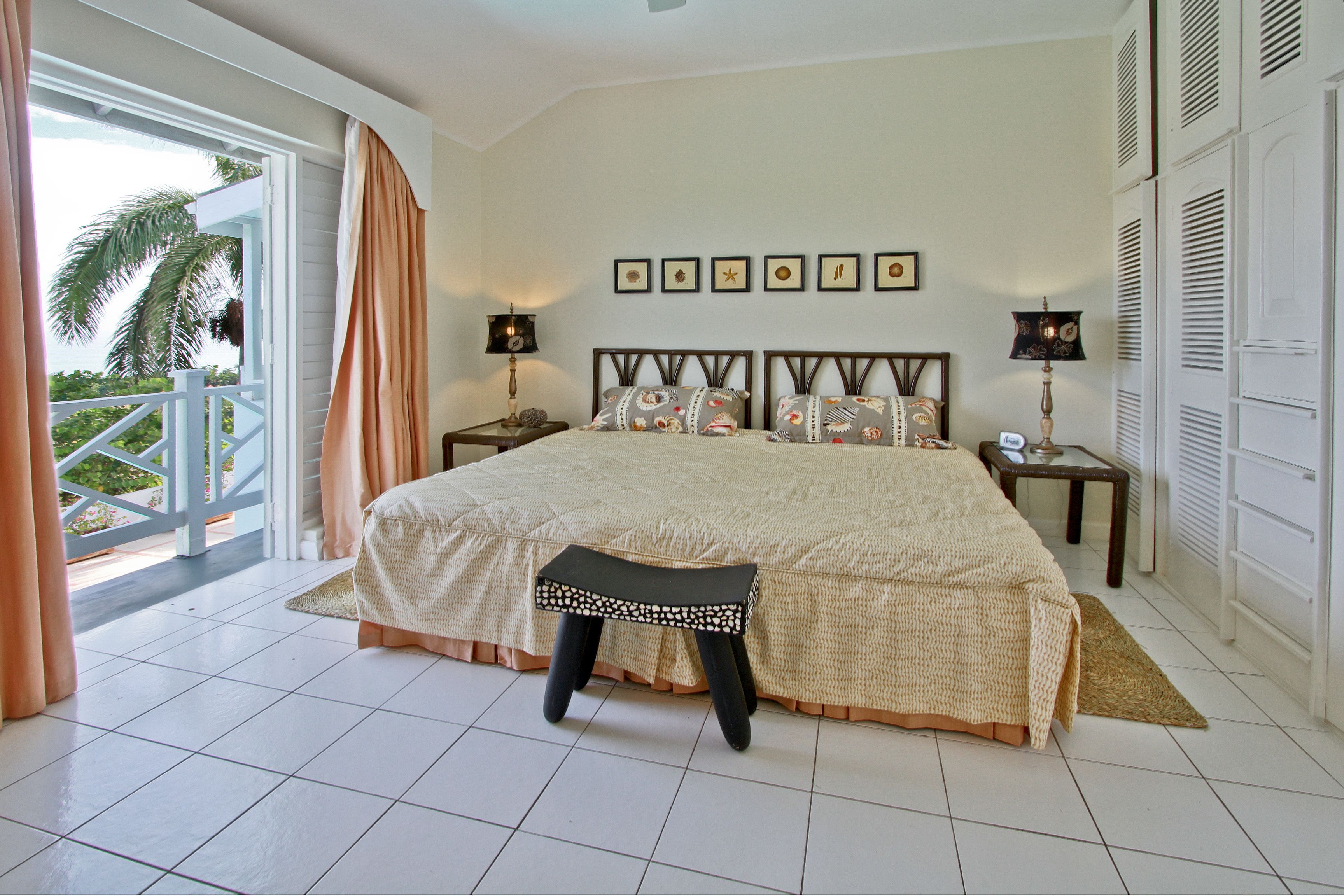 A spacious bedroom with a large bed, a small stool, two bedside tables with lamps, a view of the outdoors through a sliding door, and a series of framed pictures on the wall.