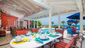 Een terras met uitzicht op de oceaan, met een witte tafel gedekt met blauwe servetten en gele placemats, omringd door rieten meubels en rode kussens.