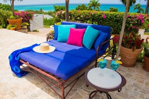 A blue sofa with colorful cushions, a hat, and two drinks on a small table, set on a patio with a view of the ocean. - Summertime, Silver Sands,4BR (Duncans)