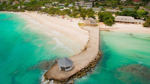 Luchtbeeld van een strandresort met een lange steiger die uitkomt bij een klein hutje, omgeven door turquoise water en een zandstrand.