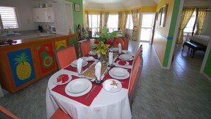 A dining area with a long table set for four, a kitchen in the background, and a bright, colorful interior. - Sol Mar, Silver Sands 3BR (Duncans)