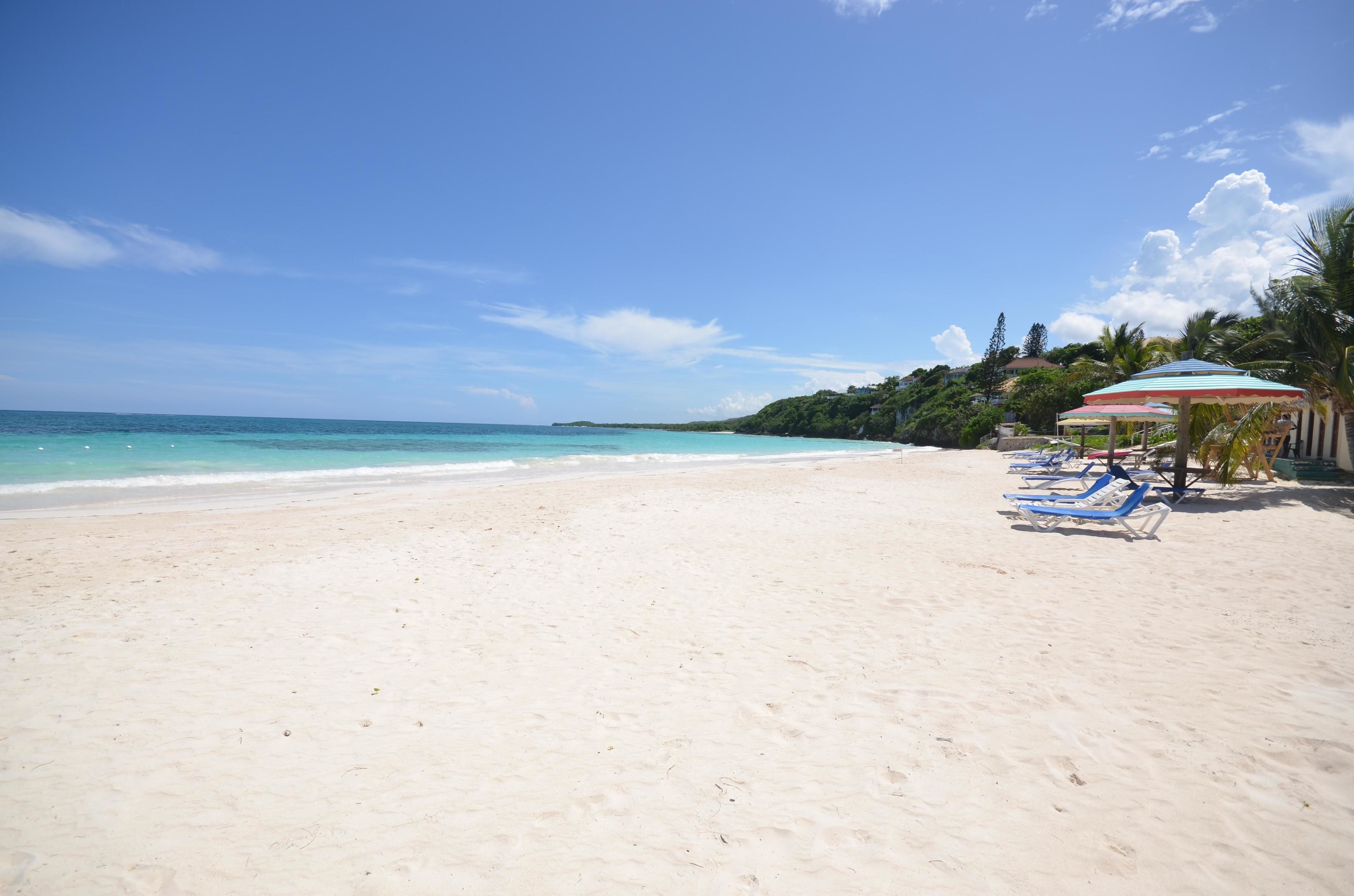 A beach with white sand, clear turquoise water, and a row of blue lounge chairs under a striped umbrella.
