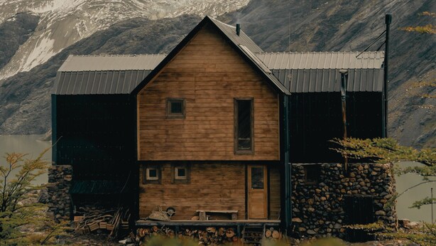 A wooden cabin with a sloped roof, situated in a mountainous area with a lake in the background. - Puesto Cagliero refugio de montaña (El Chalten)