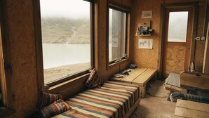 A cozy cabin room with a large window overlooking a lake, a wooden bench with cushions, and a small table with various items. - Puesto Cagliero refugio de montaña (El Chalten)