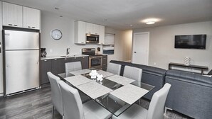 A modern kitchen with stainless steel appliances, a dining area with a glass table, and a seating area with grey upholstered sofas. - Clifton Hill Condo's (Niagara Falls)