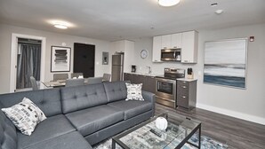 A modern living room with a grey sofa, a coffee table, and a kitchen area with stainless steel appliances. - Clifton Hill Condo's (Niagara Falls)