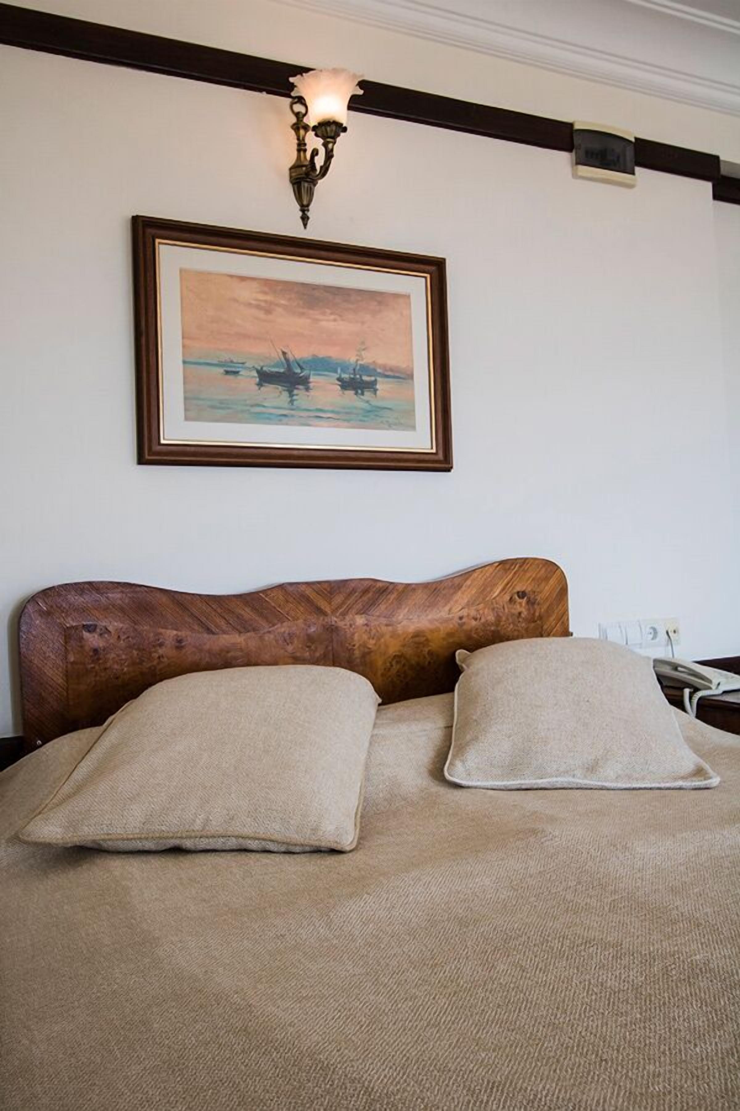 Una cama con cabecera de madera, dos almohadas color beige y un cuadro enmarcado de barcos sobre la pared.