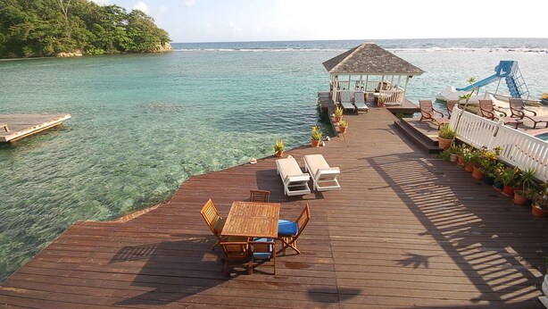 A wooden deck with lounge chairs and a table, overlooking clear turquoise water and a distant island. - San Cove, Blue Lagoon, Port Antonio 4BR (Port Antonio)