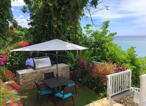 An outdoor dining area with a stone barbecue station, a table with chairs, and a large umbrella. - Scotch on the Rocks-Ocho Rios 5BR (Ocho Rios)