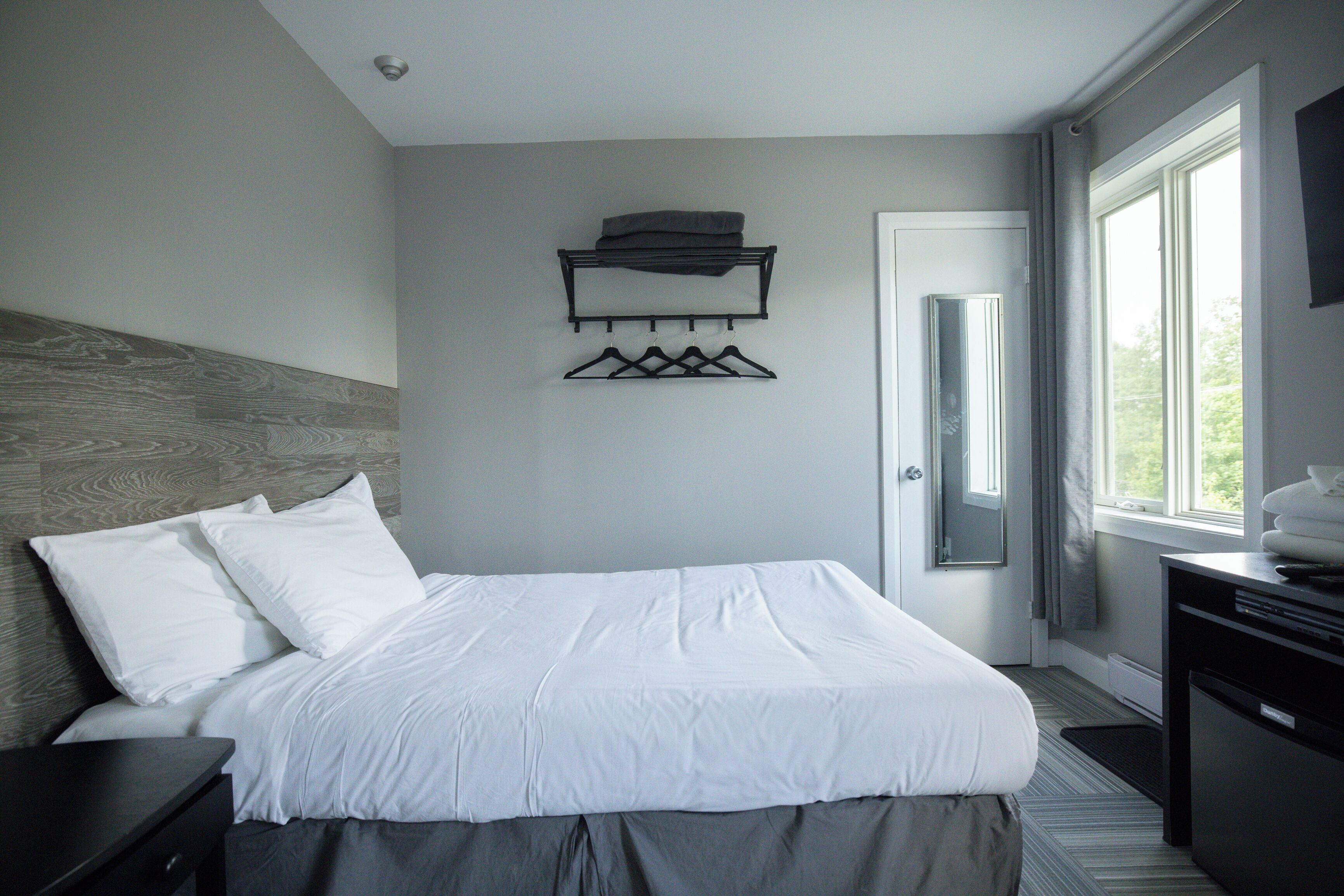 A modern hotel room with a neatly made bed, a wall-mounted coat rack, and a large window providing a view of the outdoors.