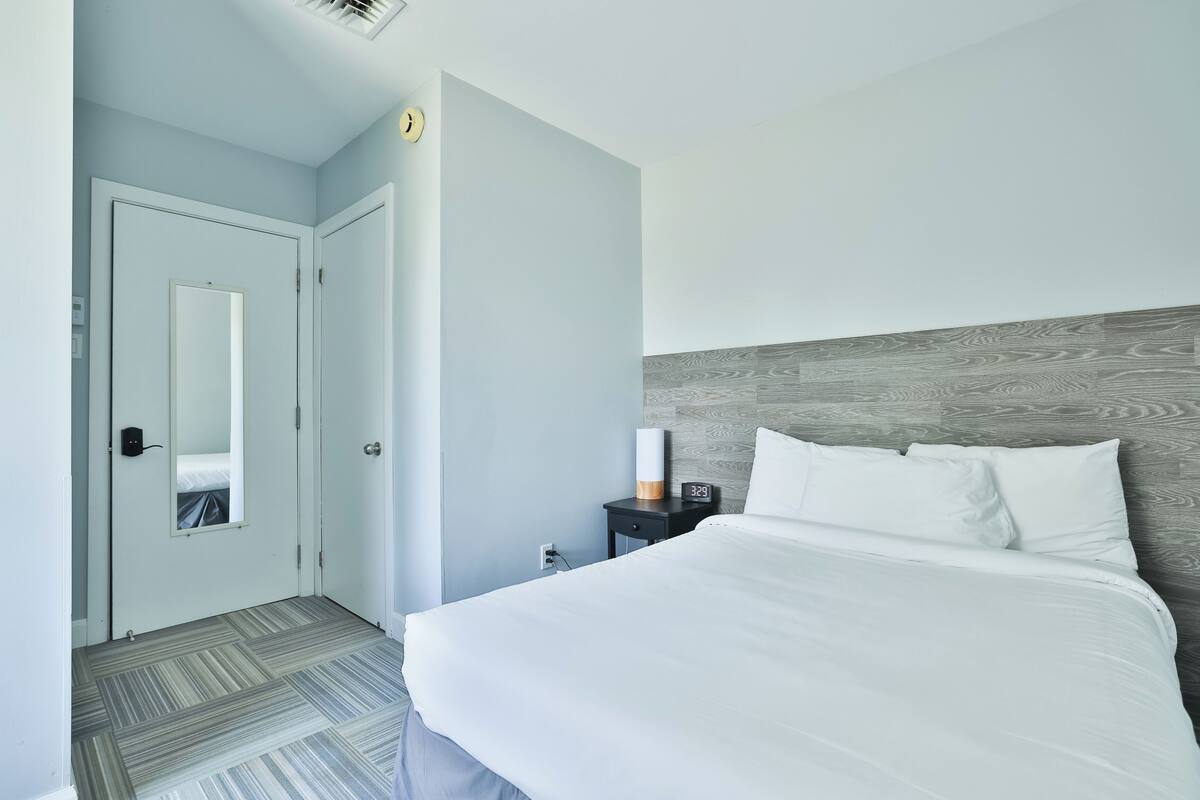 A hotel room with a bed, a nightstand, a clock, and a door.