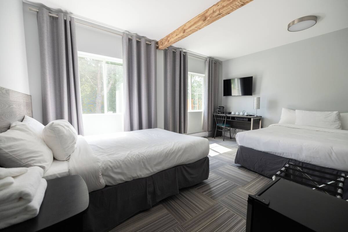 A hotel room with two beds, a desk, a TV, and large windows with curtains.