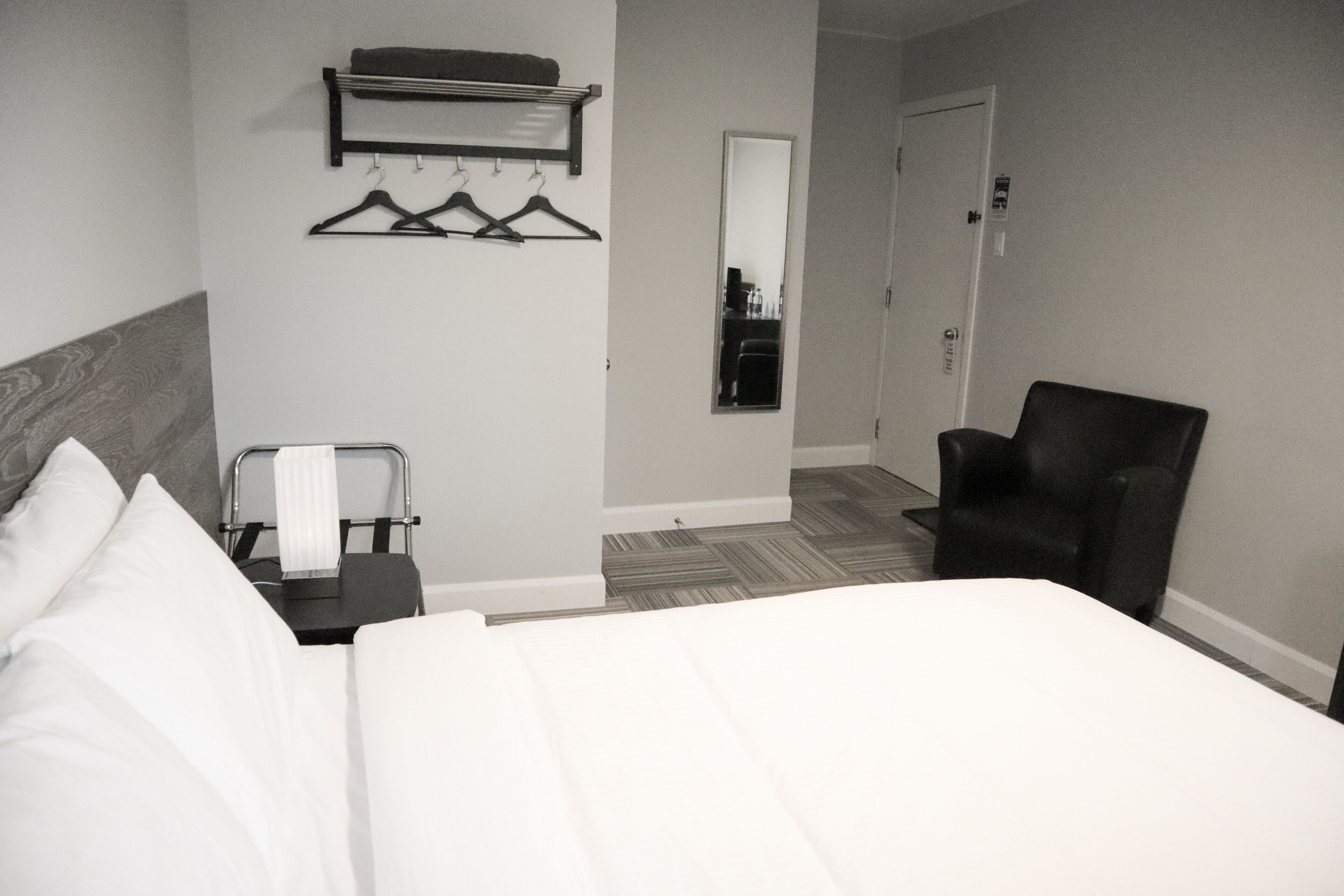 A hotel room with a bed, a chair, a mirror, and a coat rack with hangers.