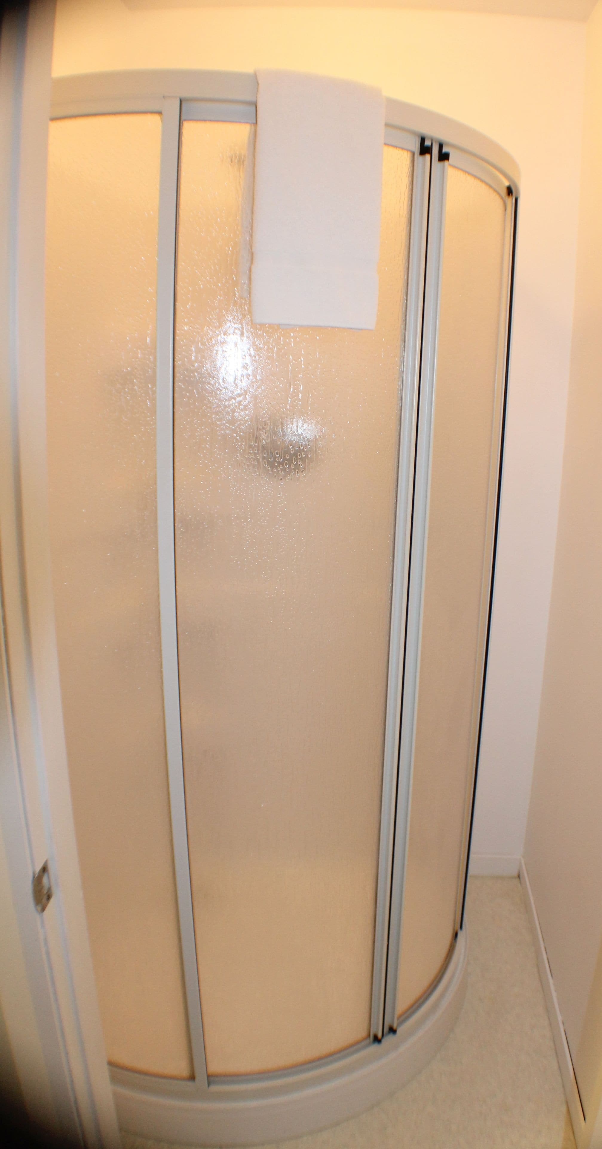 Ein Badezimmer mit Duschkabine aus Glas, einem weißen Handtuch, das an der Tür hängt, und einem beige farbenen Boden.