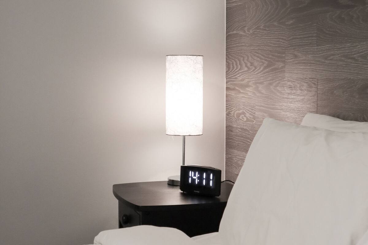A neatly made bed with white bedding, a bedside table with a lamp, and a wall with a wood-patterned headboard.