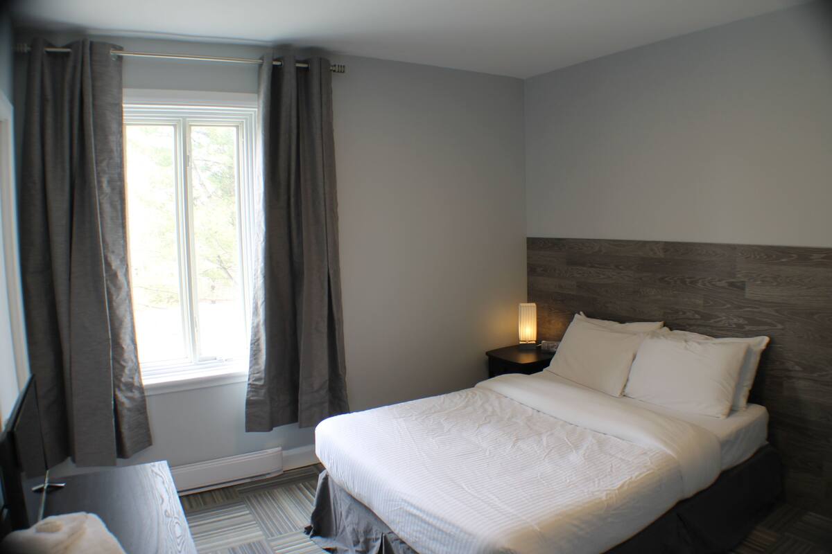 A bedroom with a bed, a nightstand with a lamp, a window with curtains, and a wooden headboard.