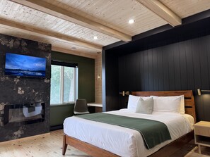 A modern bedroom with a wooden ceiling, a large bed with white bedding, a TV mounted on a dark wall, and a window with a view of trees. - Auberge Valcartier - Par Aneyro (Saint-Gabriel-de-Valcartier)