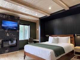 Quarto moderno com teto de madeira, uma cama grande com roupa de cama branca, uma televisão presa em uma parede escura e uma janela com vista para árvores.