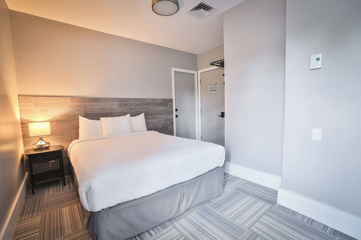 A hotel room with a large bed, a nightstand with a lamp, a wooden headboard, a door, and a thermostat on the wall.