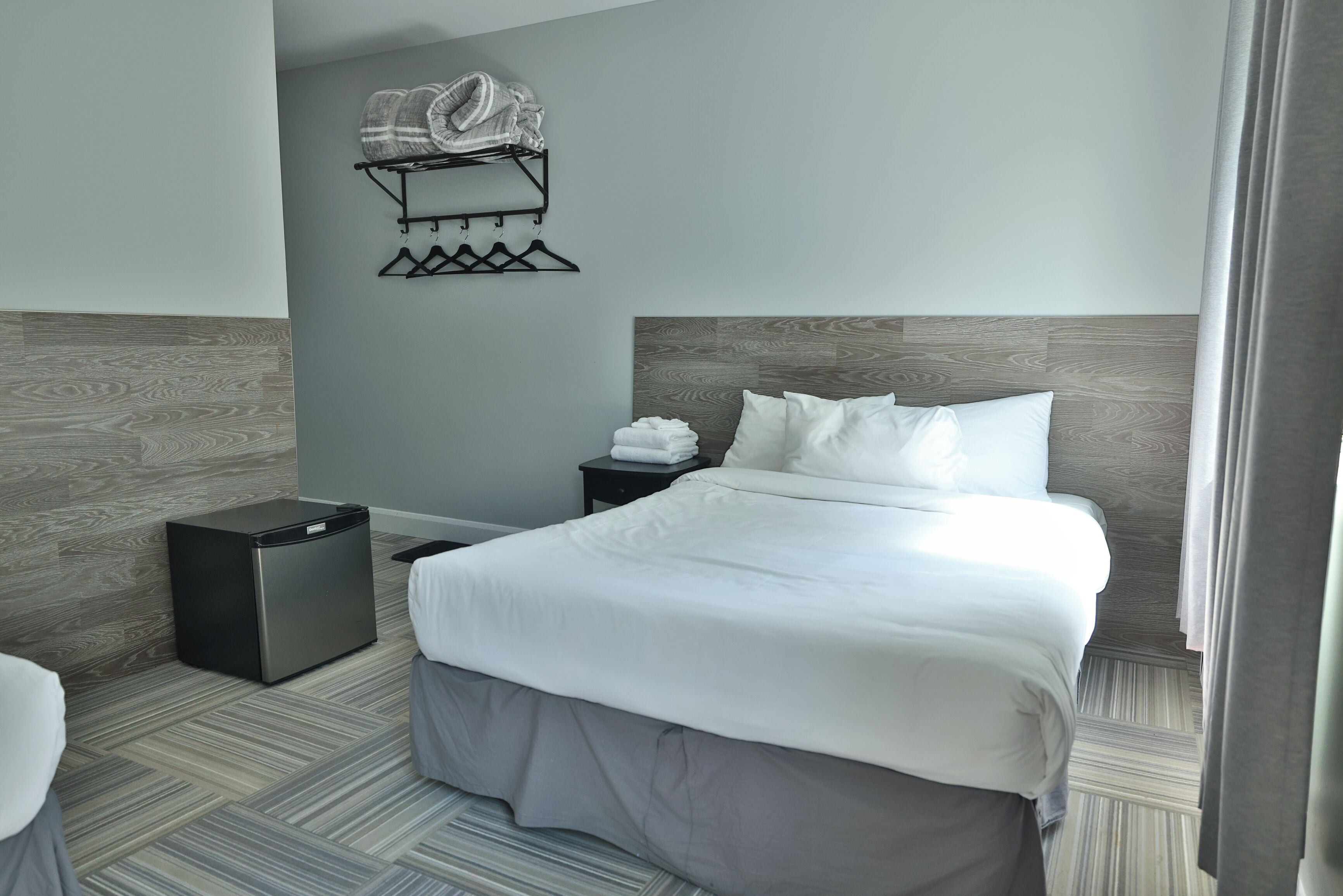 A modern hotel room with a neatly made bed, a small bedside table with a lamp, a wall-mounted towel rack, and a compact trash bin.