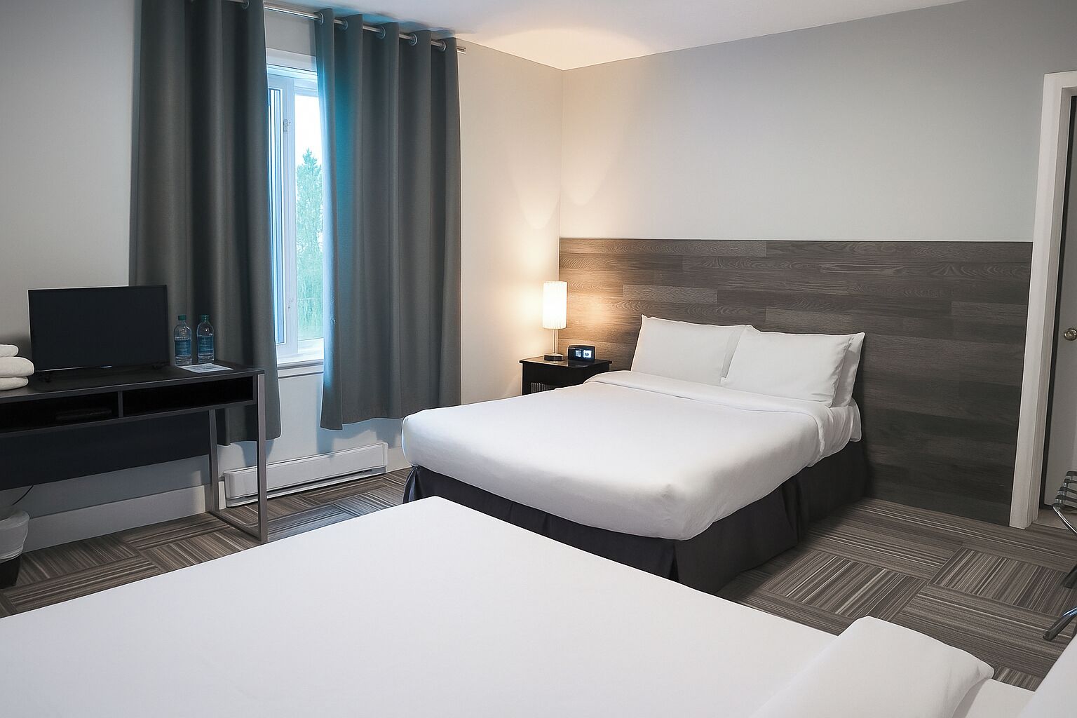A hotel room with a bed, a desk with a TV, a nightstand with a lamp, and a window with curtains.