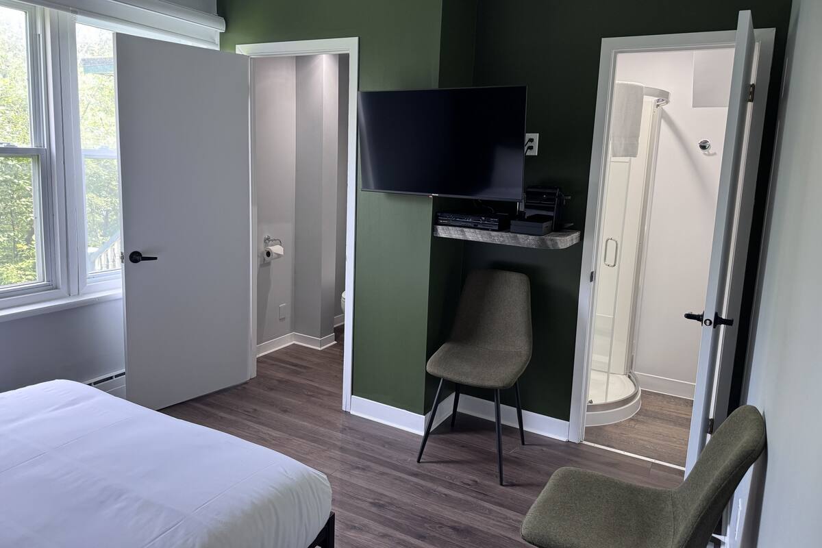 A hotel room with a bed, a TV mounted on a shelf, a chair, and a shower area.